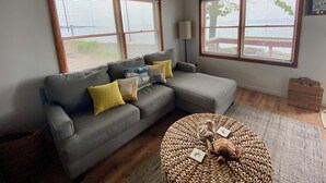 A living room with a grey sectional sofa, two windows with blinds, a lamp, a woven ottoman, and a small table with a turtle figurine. - Island Sandbar retreat with sunset views, water toys and more! Boat-Only Access (Wolcott)