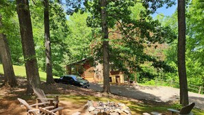 A campsite with wooden chairs and a fire pit, surrounded by trees and a cabin in the background. - Relax in Nature's Beauty: Private Log Cabin w/ two Spacious, Open Living Areas (Shipman)