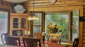 A wooden cabin interior with a dining table, chairs, a chandelier, and a large window with a view of greenery. - Relax in Nature's Beauty: Private Log Cabin w/ two Spacious, Open Living Areas (Shipman)