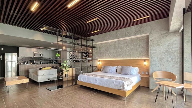Un dormitorio moderno con una cama grande, un sofá, una mesa de comedor y una zona de cocina visible a través de un separador de vidrio.