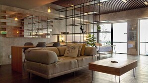 Una sala moderna con sofá, mesa de centro y un escritorio de madera. Se observa una escalera con barandilla y una planta en el alféizar.