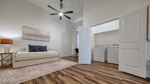 A bedroom with a bed, a nightstand, a lamp, a ceiling fan, a painting, and a walk-in closet with a washing machine. - Brand New, Beautifully Appointed, Country Cottage! (Lindale)
