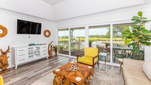 A spacious living room with a wooden coffee table, a yellow armchair, and a white cabinet with glass doors. A flat-screen TV is mounted on the wall, and there is a large window offering a view of the outdoors. - Golden Isles Getaway -**ASK ABOUT SNOWBIRD SPECIALS** (Brunswick)