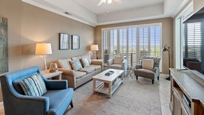 A living room with a beige sofa, blue armchair, and a coffee table. There are two lamps, a ceiling fan, and a TV on a wooden stand. - Wharf 703|Waterfront View|2 bedroom|Pool|sleeps 8! (Orange Beach)