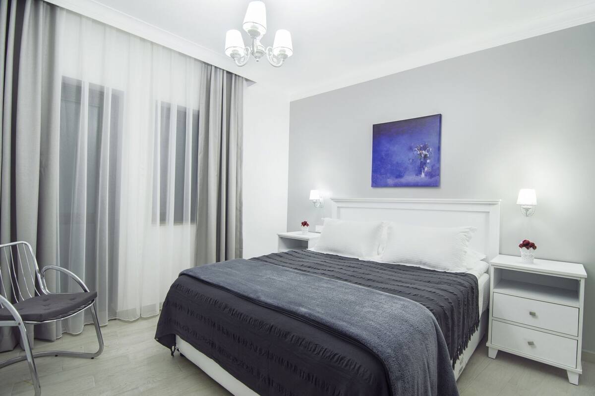 A modern hotel room with a large bed, a bedside table with a lamp, a chair, and a painting on the wall.