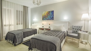 A hotel room with two beds, a chair, a small table, a lamp, and a painting on the wall. - Rodina Suites (Kusadasi)