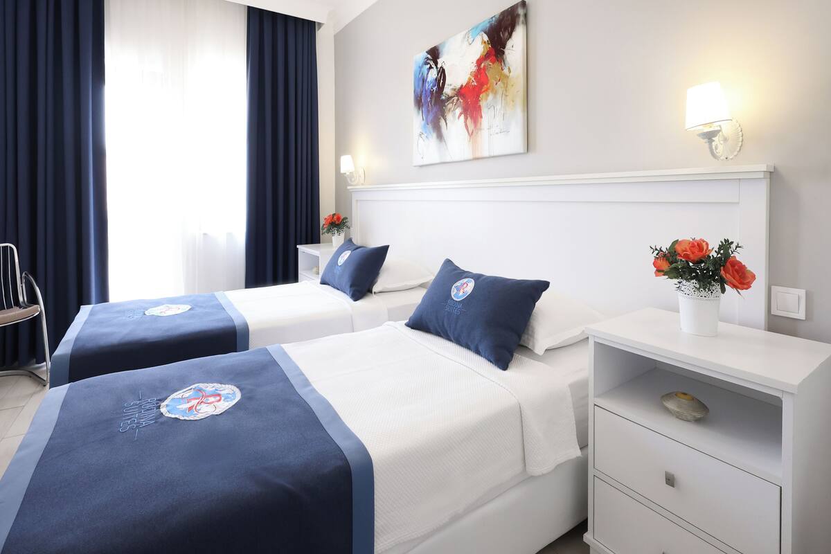A hotel room with two beds, a nightstand, a chair, a painting, and a window with curtains.