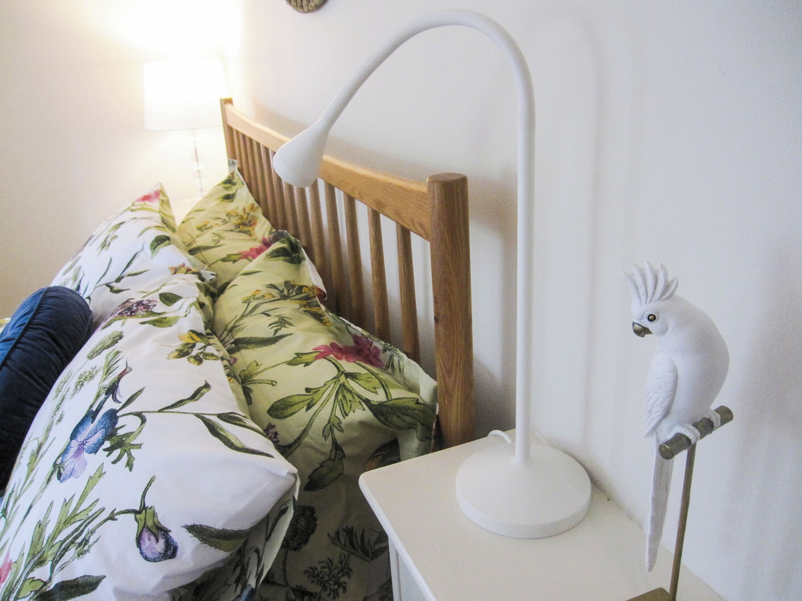 Un letto con biancheria a fiori, una lampada da comodino bianca e una statuetta di pappagallo bianco su un piedistallo.