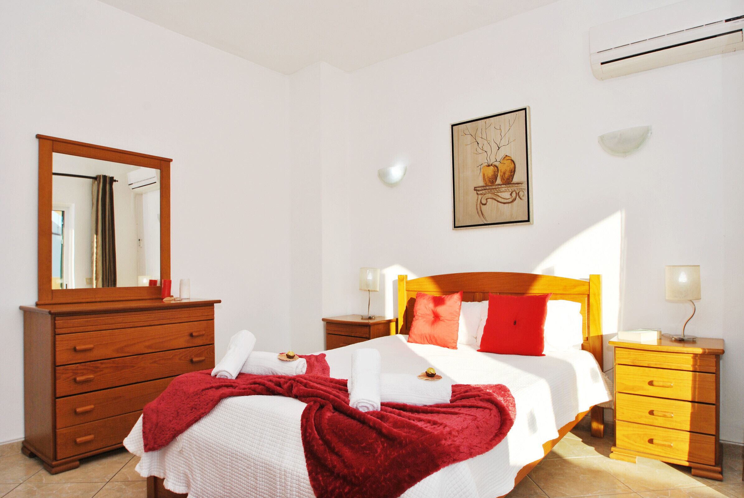 Un lit bien fait, avec du linge de lit blanc et rouge, une commode en bois, une table de chevet avec une lampe et un climatiseur fixé au mur.