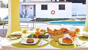 A breakfast setup with a pool view, including plates of food, drinks, and a basket of bread. - Traditional villa with BBQ and heatable pool (Albufeira)