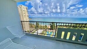 A balcony with two white metal chairs, a view of the ocean, a beach, and a tall building in the background. - Caribbean Beach front King bed with large balcony (Luquillo)