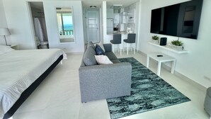 A modern hotel room with a large bed, a grey sofa, a small kitchen area, and a TV mounted on the wall. - Caribbean Beach front King bed with large balcony (Luquillo)