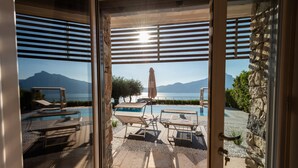 A pool area with lounge chairs and an umbrella, a stone wall, and a view of mountains and water. - Luxury Villa Vela (Torri del Benaco)