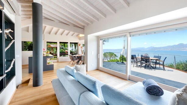 A modern interior with a large window, a sofa, and a dining area with outdoor seating. - Luxury Villa Vela (Torri del Benaco)