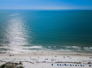 An aerial view of a beach with clear skies, calm waters, and a sandy shore with several people and blue lounge chairs.