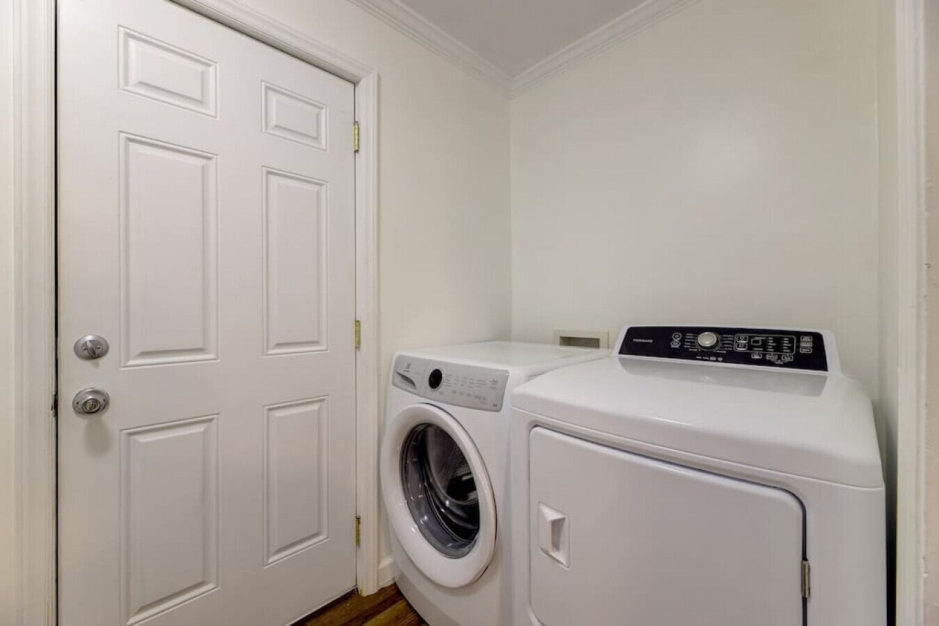 Une buanderie équipée d’un lave-linge et d’un sèche-linge à chargement frontal, d’une porte blanche et d’une petite prise électrique au mur.