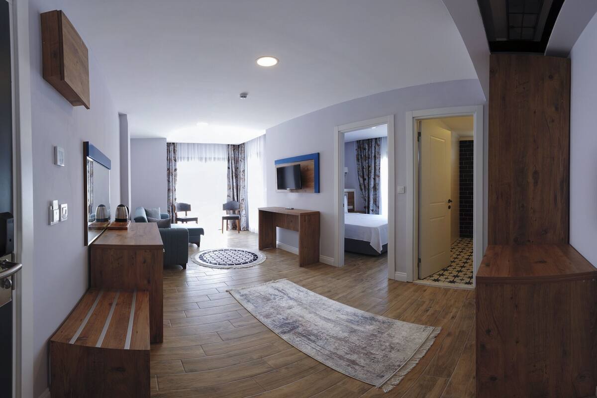 A modern hotel room with a wooden bench, a TV, a desk, a bed, and a view of the outside.