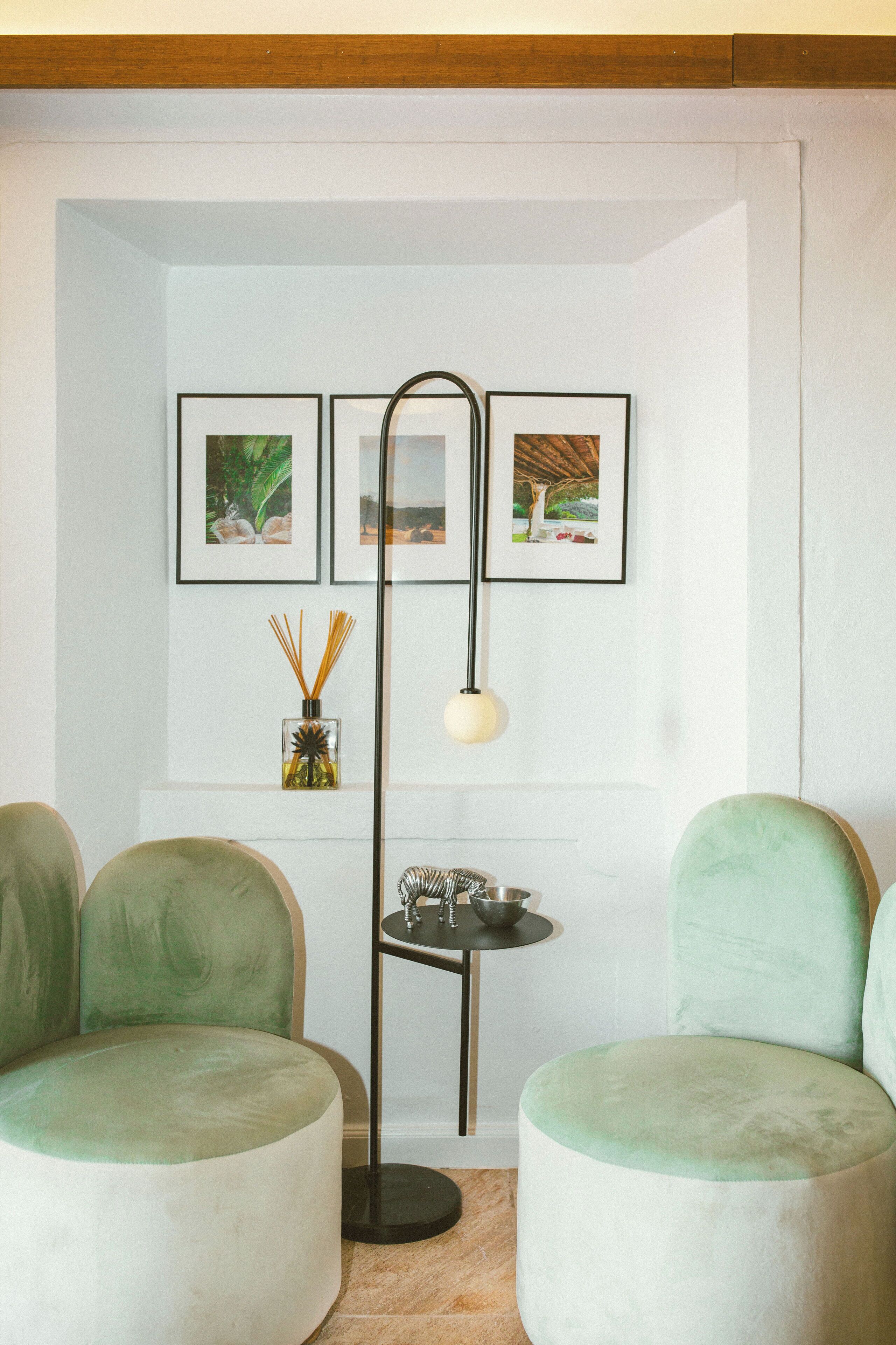 Une pièce comprenant deux fauteuils verts, un lampadaire, une petite table avec un chandelier et trois tableaux encadrés accrochés au mur.
