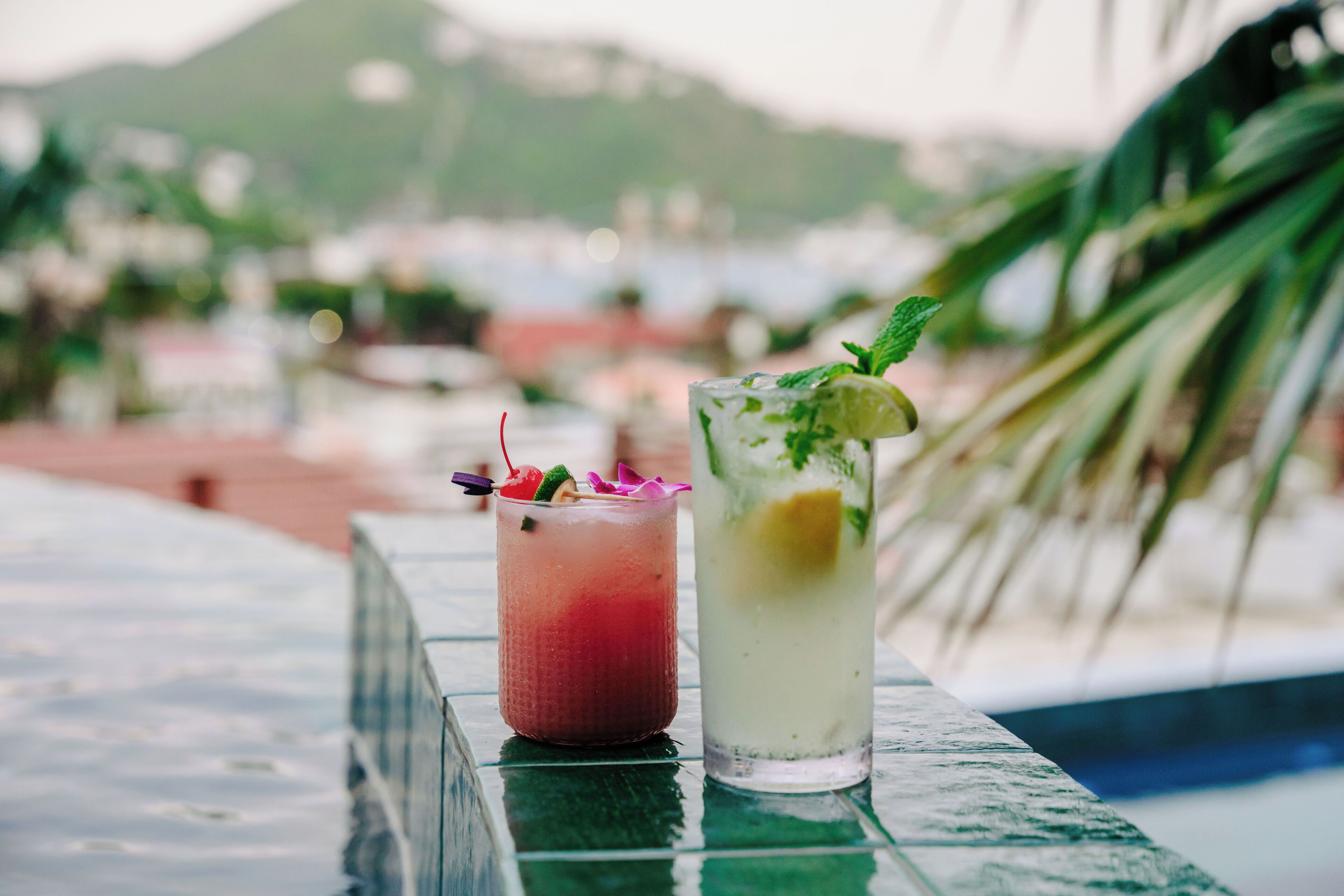 Two cocktails on a table with a pool and mountains in the background.
