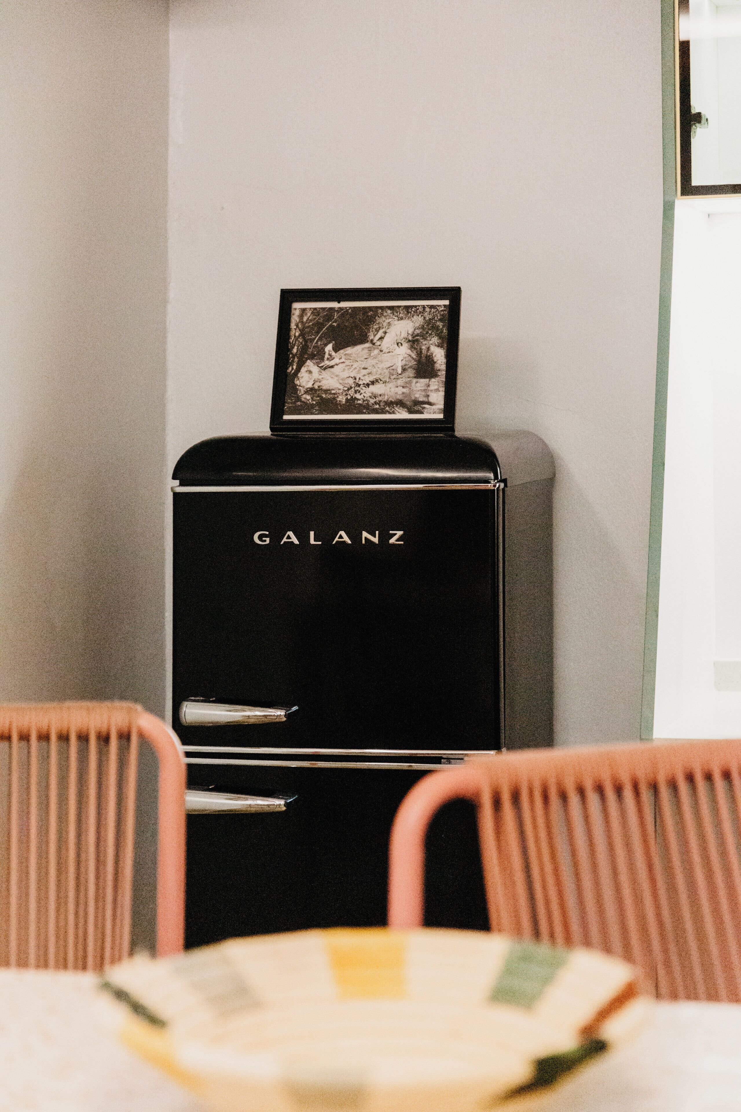 Un réfrigérateur Galanz noir, orné d’une image sur le dessus, deux chaises roses et une table avec un set de table blanc et jaune.