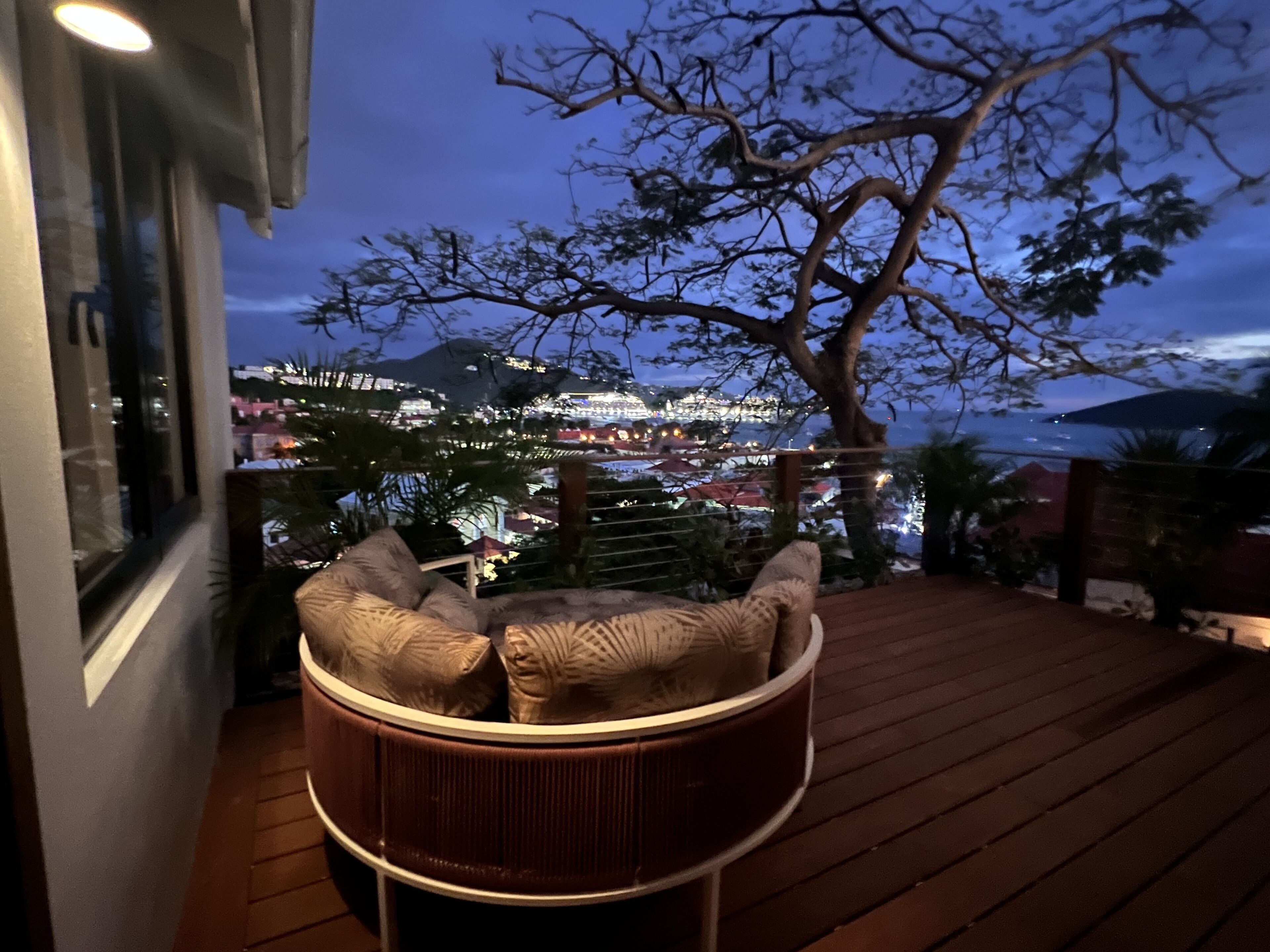 A wooden deck with a wicker chair and cushions, overlooking a cityscape at dusk.