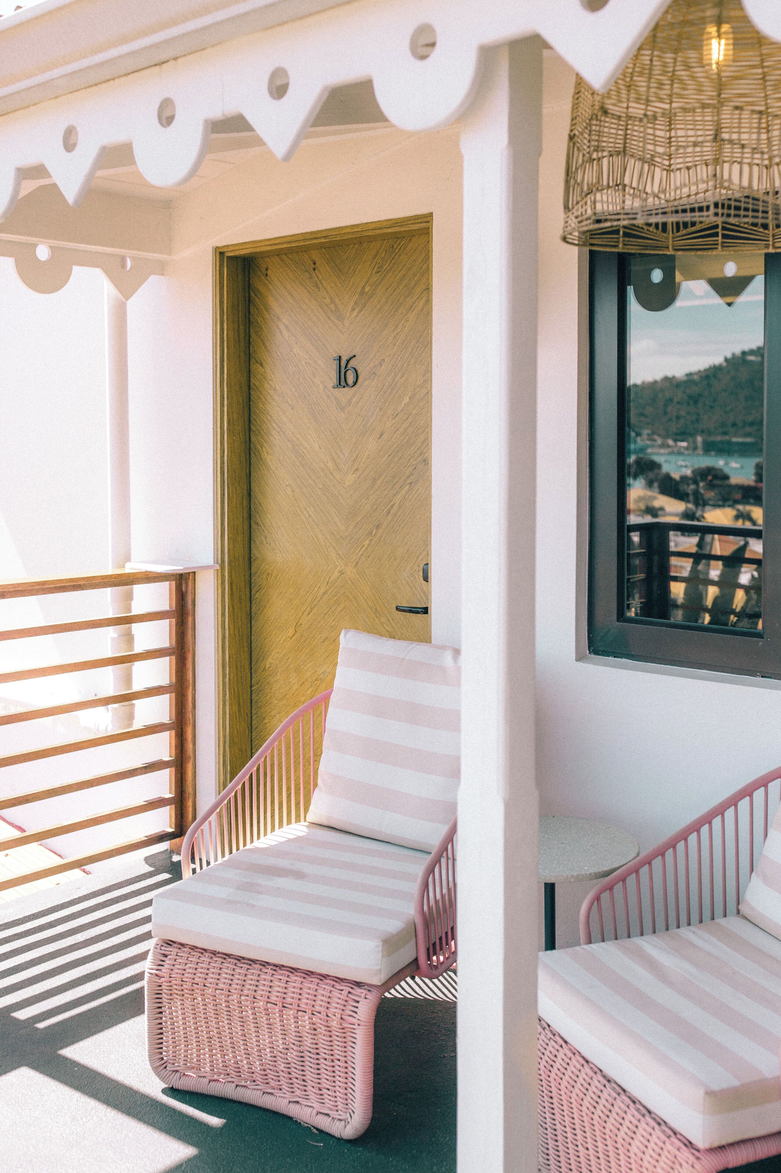 A balcony with wicker lounge chairs, a wooden door with the number 16, and a view of a coastal landscape through a window.