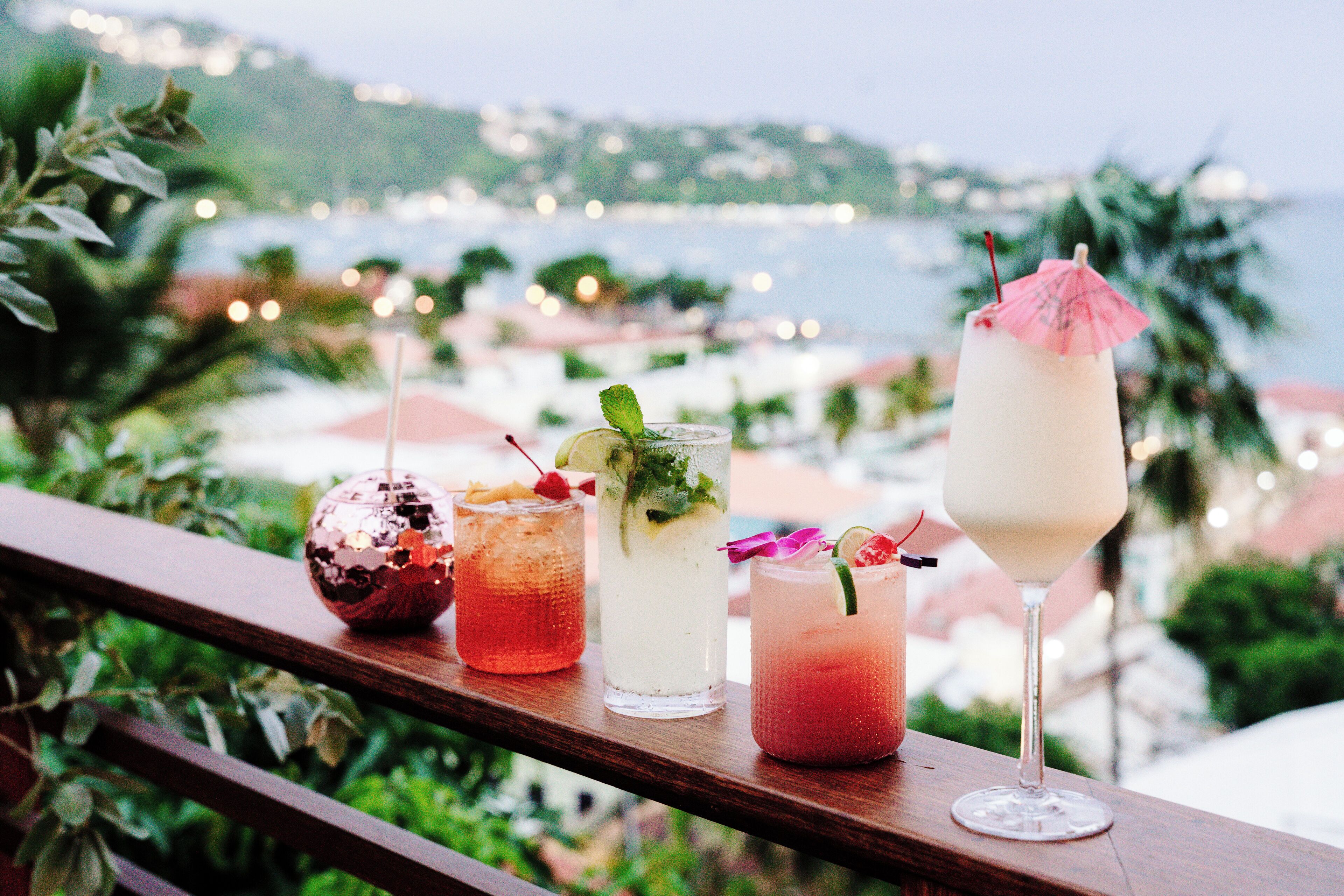 A wooden railing with three drinks on it, overlooking a coastal area with buildings and greenery.