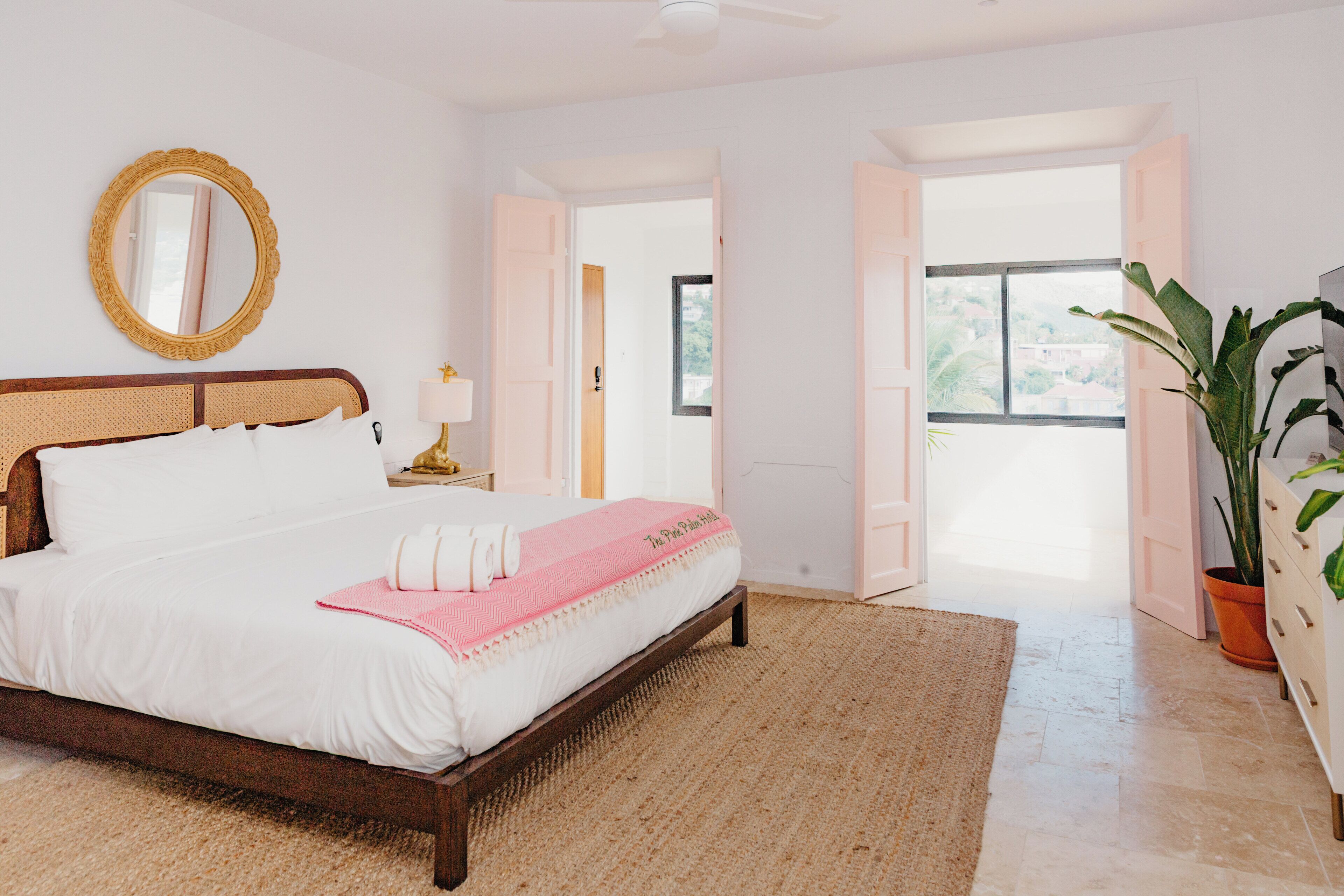A bedroom with a bed, a mirror, a nightstand, a potted plant, and a view of the outdoors.
