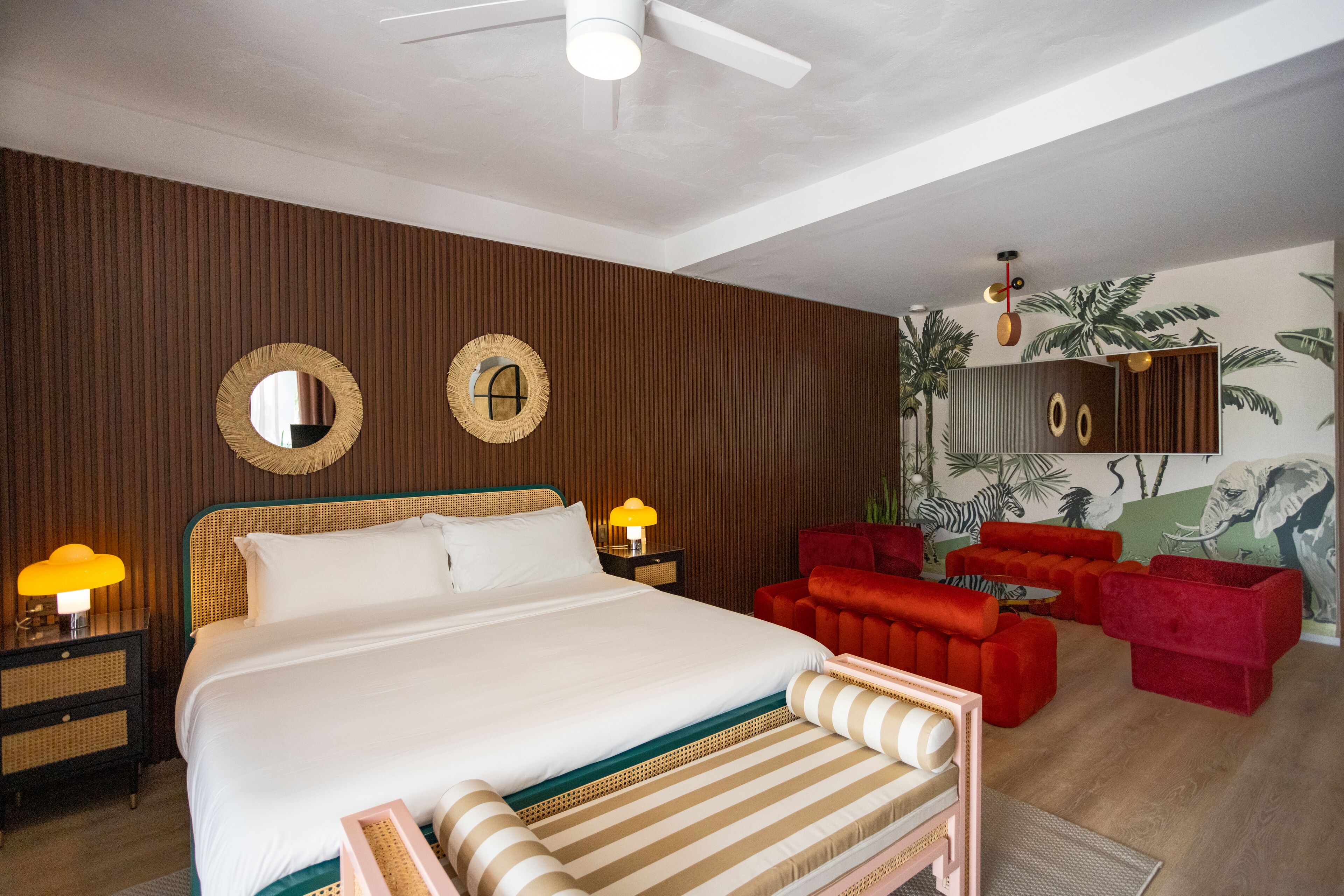 Quarto de hotel com cama grande, ventoinha no teto, sofá vermelho e um mural na parede.
