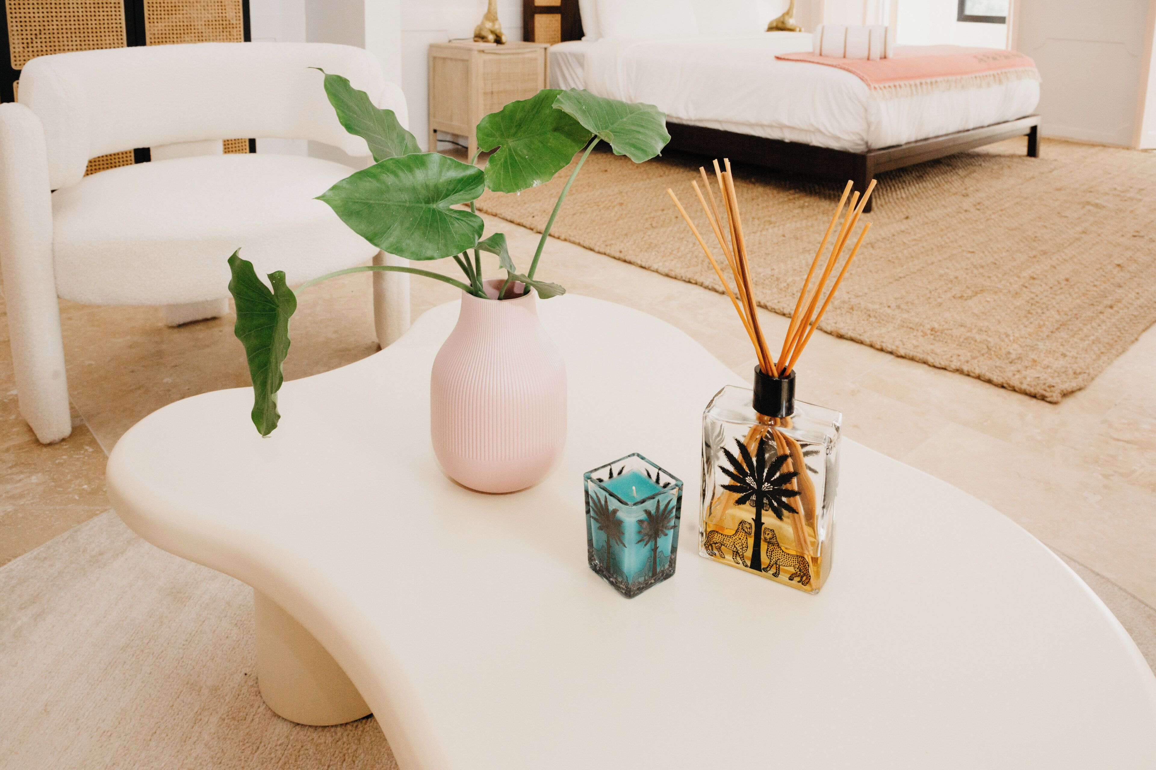 Ein modernes Schlafzimmer mit einem weißen Bett, einem weißen Sessel, einer großen Grünpflanze, einer rosa Vase und einer Glasflasche mit Schilfrohrstäbchen.