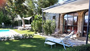 Un espace aménagé au bord de la piscine, avec un bâtiment en pierre, une terrasse couverte et des chaises longues.