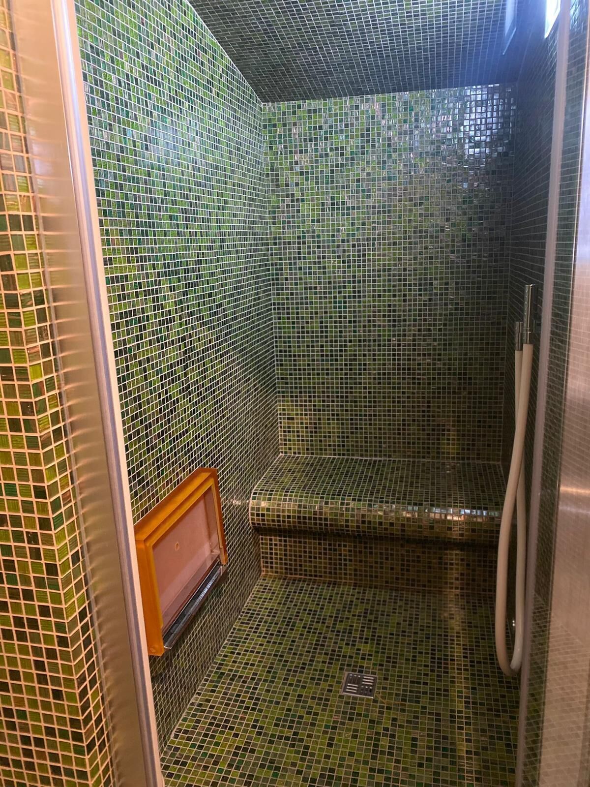 Ducha con mosaicos verdes, un banco de madera y un cabezal de ducha manual.