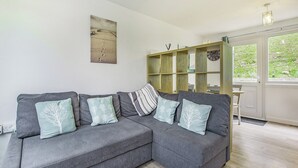 A grey sectional sofa with tree-patterned cushions, a wooden bookshelf, and a large window with a view of greenery. - 2 bedroom accommodation in Freshwater East, near Pembroke (Freshwater East, near Pembroke)