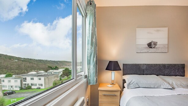 A hotel room with a bed, a bedside table with a lamp, a dresser, and a window with a view of buildings and hills. - 2 bedroom accommodation in Freshwater East, near Pembroke (Freshwater East, near Pembroke)