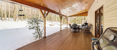 A covered wooden deck with a grill and outdoor seating area, overlooking a snowy landscape.
