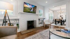 A modern living room with a fireplace, a large flat-screen TV, and a dining area with a chandelier. - Location!! - The Davidson - Charming Home in Fayetteville Historic District (Fayetteville)