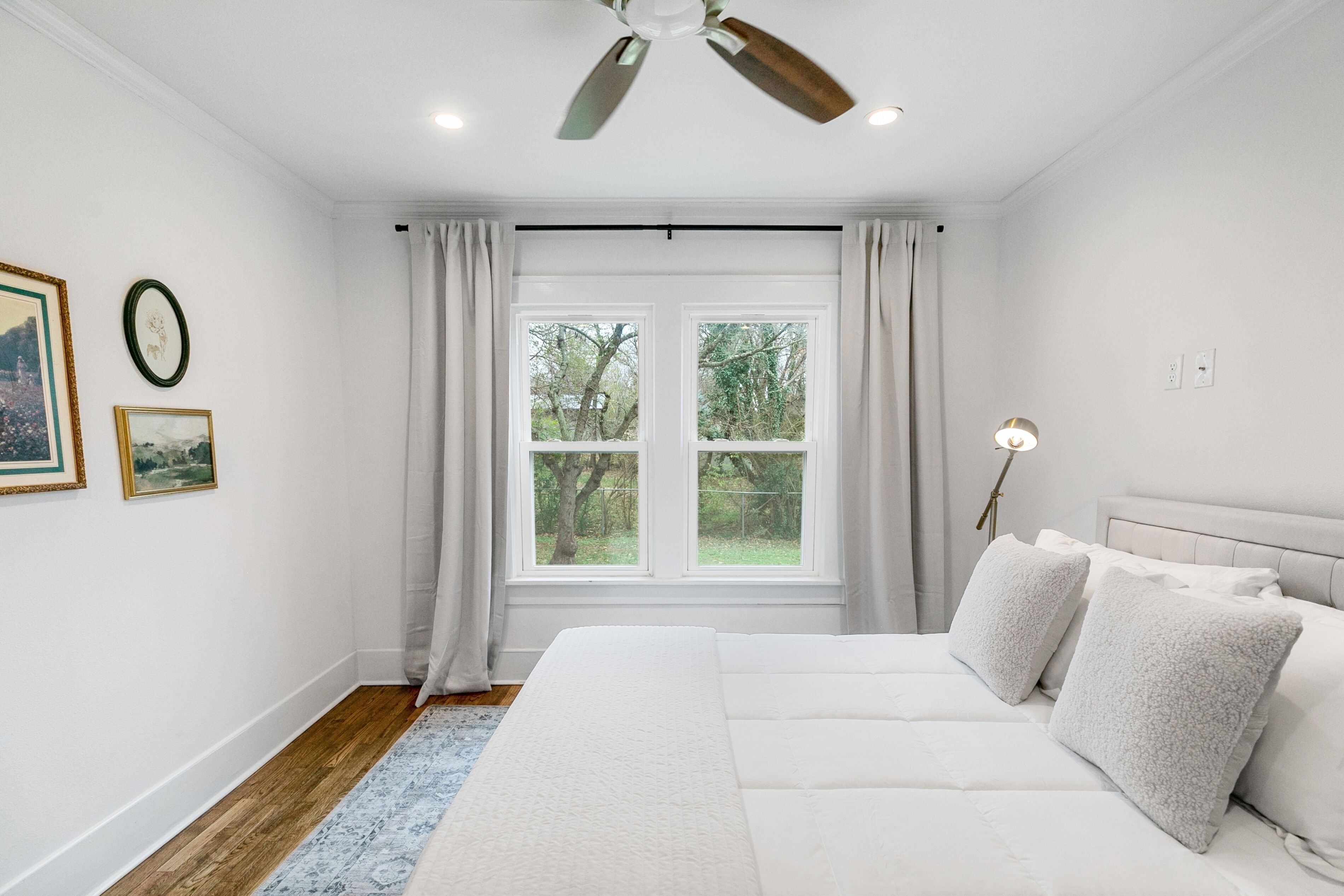 Une chambre avec un grand lit, un ventilateur de plafond, une fenêtre avec des rideaux et des tableaux encadrés accrochés au mur.