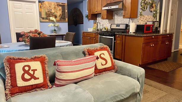 Una sala de estar con un sofá gris, dos cojines decorativos, un comedor con mesa y sillas, y una cocina con armarios de madera y electrodomésticos.