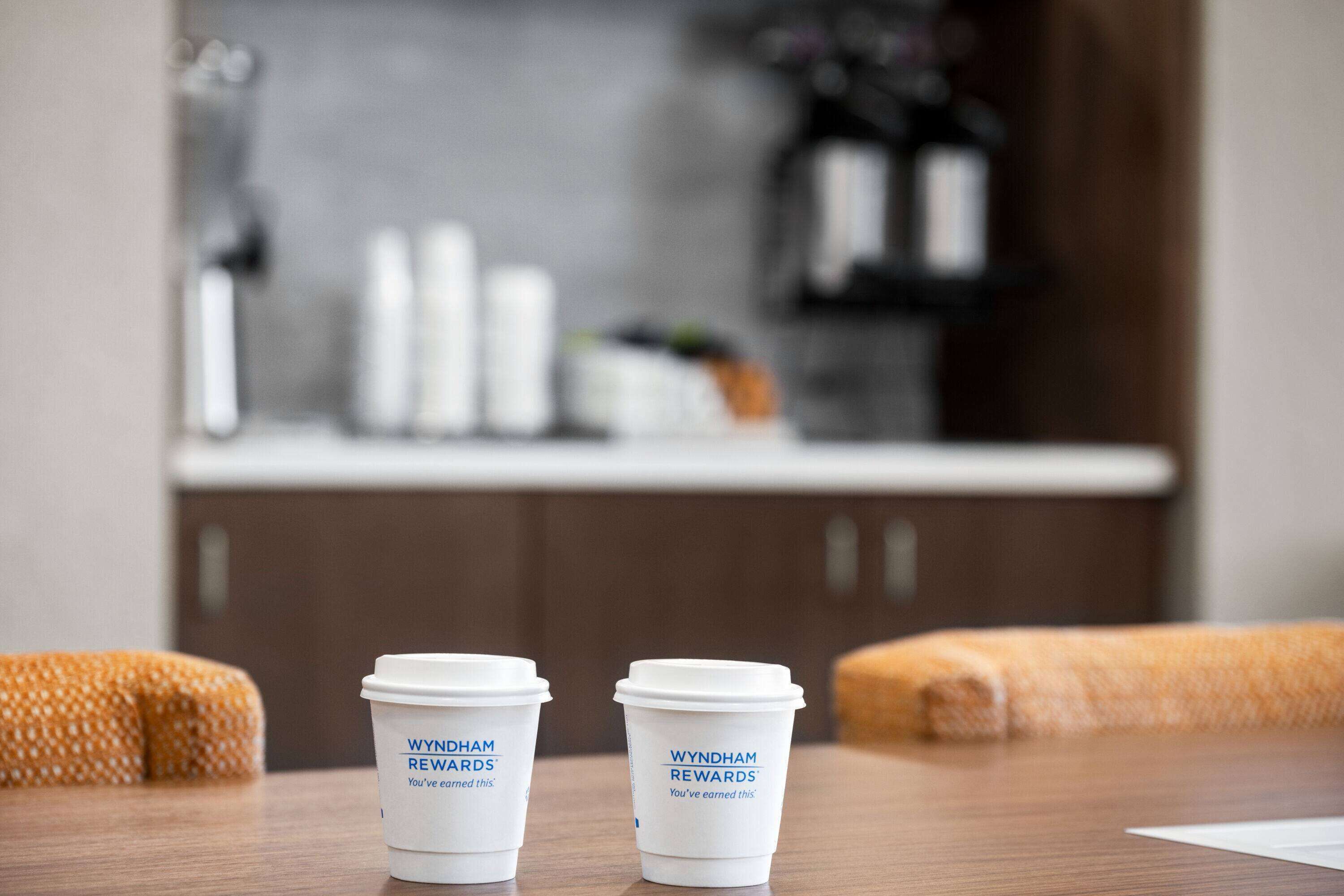 Deux tasses à café portant le logo « Wyndham Rewards » sont posées sur une table en bois dans une pièce où l’on aperçoit une cuisine en arrière-plan.