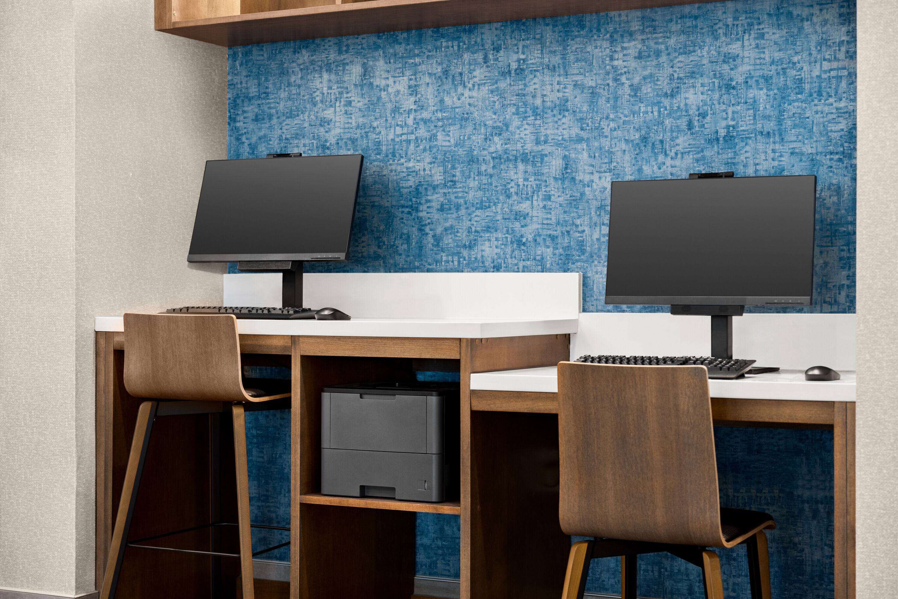 A modern office space with two desks, each with a computer monitor, keyboard, and mouse. There are two chairs, and a printer is visible on the left desk.