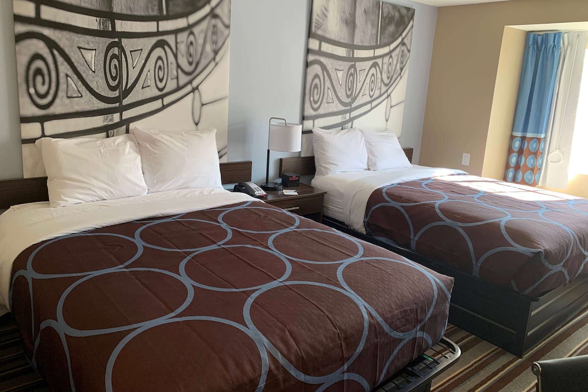Een hotelkamer met twee bedden, elk voorzien van een decoratieve spreidek, een nachtkastje met een lamp, en een groot abstract kunstwerk aan de muur.