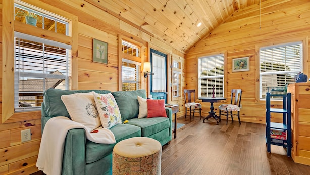 Interior acogedor de una cabaña con un sofá verde, un taburete estampado, paredes de madera y una zona de comedor con mesa y sillas.