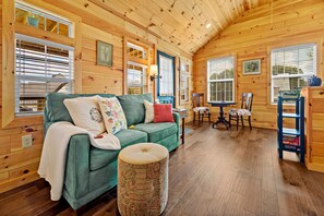 L’intérieur d’un chalet accueillant, avec un canapé vert, un pouf à motifs, des murs en bois et un coin repas comprenant une table et des chaises.