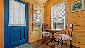 Interior de una cabaña de madera con una puerta azul, dos ventanas con persianas, una mesa de comedor con sillas y un cuadro en la pared.