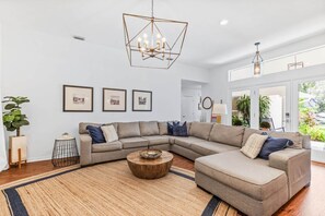 A modern living room with a sectional sofa, a wooden coffee table, and a geometric chandelier. - The Water's Edge, St. Augustine (Vilano Beach)