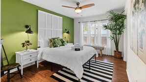 Un dormitorio con cama, mesitas de noche, ventilador de techo, ventana con cortinas, una planta en maceta y una alfombra a rayas.