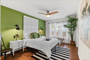 A bedroom with a bed, bedside tables, a ceiling fan, a window with curtains, a potted plant, and a striped rug. - The Water's Edge, St. Augustine (Vilano Beach)
