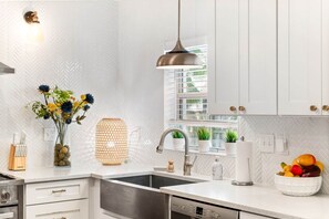 Cozinha moderna com armários brancos, pia de aço inoxidável, luminária pendente e janela com persianas.