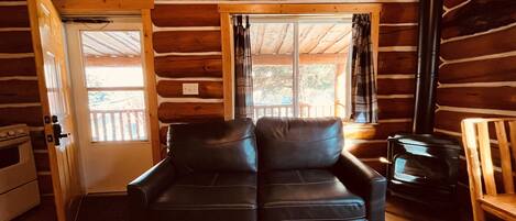 L’intérieur d’une cabane en rondins, avec un canapé en cuir noir, une chaise en bois, une cheminée et une porte donnant sur l’extérieur.
