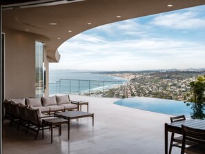 A modern balcony with a curved glass railing overlooking a coastal cityscape and a clear blue sky. - La Jolla villa with 5 bedrooms, private pool, hot tub, and ocean views. (La Jolla)